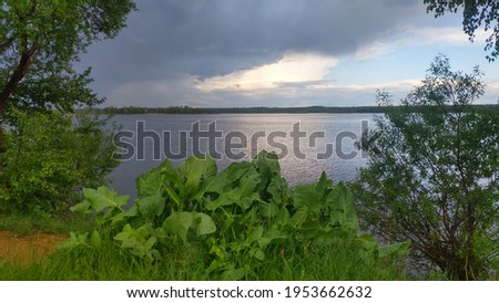 A summer thunderstorm is coming to the lake, large storm clouds are gathering. View of the lake from the shore among the bushes and trees.