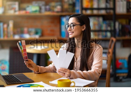 Young creative female graphic designer with color sets at cafe, using laptop and drinking coffee, empty space. Happy woman looking at colorful palette, working on computer ddistantly