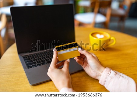Online shopping in internet concept. Woman using credit card and laptop while sitting at cafe and drinking coffee, computer with black blank screen. Mockup for advertisement