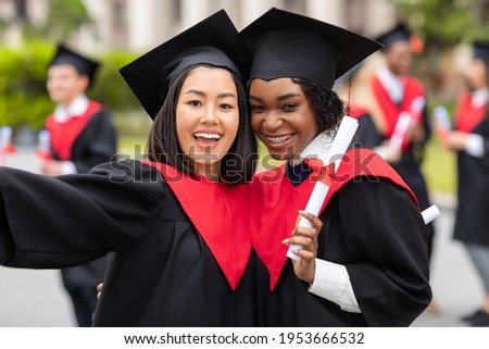 Two cheerful young ladies asian and african american girlfriends in graduation robes taking selfie, showing diplomas and smiling at camera, closeup portrait over university campus