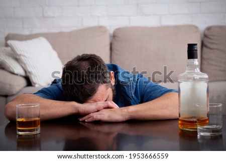 Alcoholism, addiction, weakness and drinking whiskey alone. Sad young guy in depression lies on table with bottle and glass of alcohol and suffers from sadness, in living room interior, free space Royalty-Free Stock Photo #1953666559