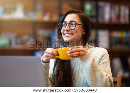 Benefits of remote work. Dreamy lady enjoying coffee during work on laptop computer in city cafe, smiling and looking aside. Young freelancer having break while working online in internet Royalty-Free Stock Photo #1953680449