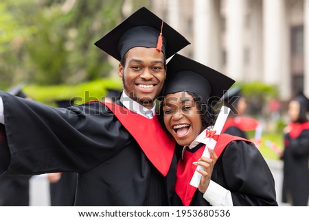 Happy african american couple students in graduation dresses and hats taking selfie together, posing at university campus, enjoying and celebrating graduation, closeup portrait Royalty-Free Stock Photo #1953680536