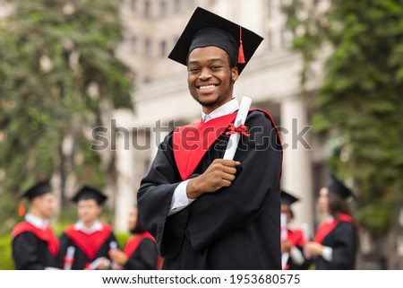 Cheerful african american guy in graduation costume showing his diploma and smiling at camera, black male student posing over international group of students at university campus, copy space Royalty-Free Stock Photo #1953680575