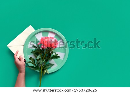 Hands holding tray with single pink peony flower over paper envelope on green background. Trendy casual natural eco friendly flat lay with copy-space. Summer birthday or Mother's day greeting card