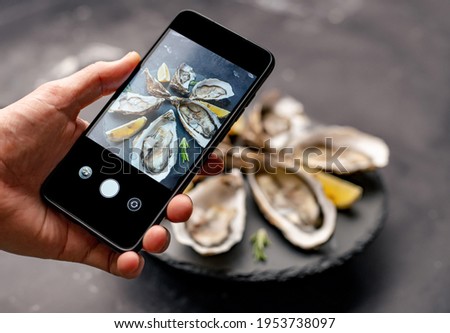 Woman holding smartphone and taking picture of fresh oysters with lemon on black plate