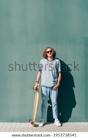 vertical portrait of a caucasian young skateboarder looking at camera. He is wearing casual clothes and sunglasses. Green wall background