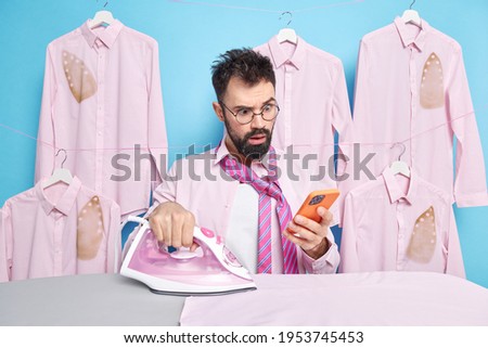 Shocked busy businessman checks newsfeed via mobile phone while ironing clothes uses steam electric iron being inattentive and burned many shirts wears round transparent spectacles formal outfit.