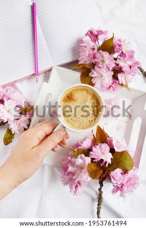 Big cup of coffee with milk and notebook on the white bedsheet. Sakura flowers around cut of hot coffee. Top view. Spring time. Human hand is taking cup of coffee. Work from home or anywhere. Cozy.
