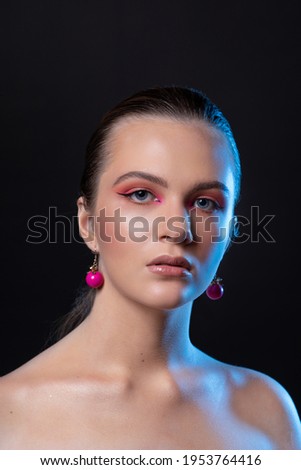 High beauty photo of a lovely young girl with wonderful professional makeup, round pink earrings, with long brown hair. Posing over black background. Close-up. Studio shot. Portrait
