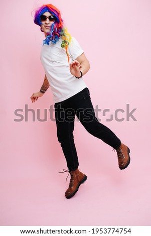 Feminine man playing girl, jumping around and playing cute. He is wearing rainbow colored wig and sunglasses. Over pink background