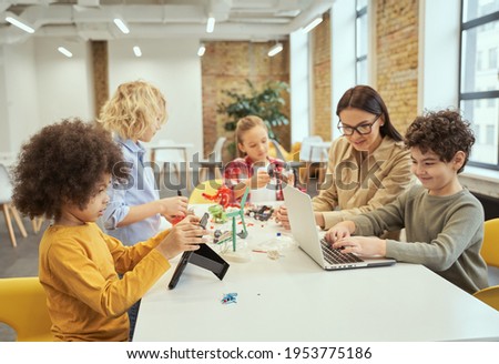 Group of diverse kids working together with young female teacher, using digital devices and playing with mechanical toys while sitting at the table during STEM class Royalty-Free Stock Photo #1953775186