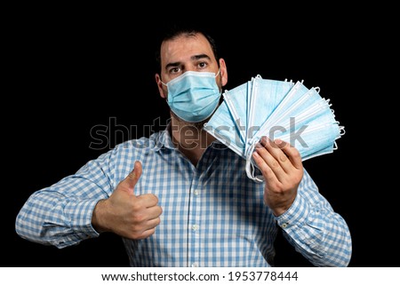 Handsome bearded man wearing a plaid shirt and surgical mask to protect himself from coronavirus by lifting his thumb up with one hand and holding several surgical masks with the other hand.