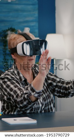 Retired woman experiencing virtual reality using vr headset in living room working from home. Old remote employee searching, analysing financial reports while elderly wife watching tv in background