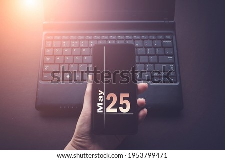 May 25th. Day 25 of month, Calendar date. Hand Holding Mobile Phone on Laptop Computer dark background with sunshine.  Spring month, day of the year concept
