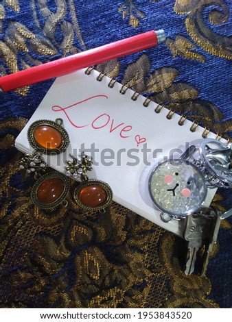 This isa picture of love on a diary, with red pen and with red heart and their is also a kitty Keychain with it and brown jewelry beside it and it shows luv