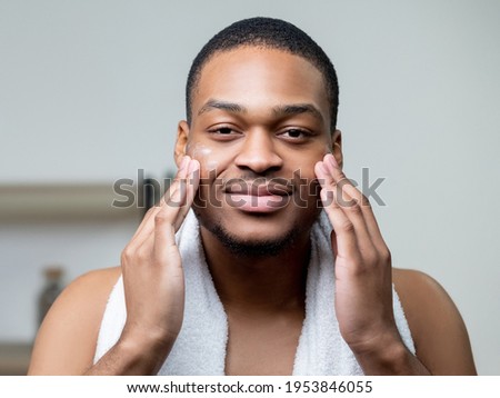 Man facial care. Skin moisturizing. Acne treatment. Confident satisfied African guy applying cream on dark smooth face with hands on light background. Royalty-Free Stock Photo #1953846055