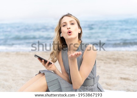 Joyful blonde girl sending a kiss gesture holding a mobile phone and wearing headphones sitting on a sand near the ocean 