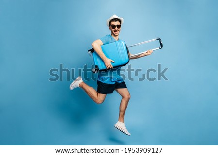 Full-length photo of a happy caucasian amazed man in a sunglasses and hat, in casual wear, jumping with a suitcase in his hands on a blue background. Excited about long-awaited vacation, smiling Royalty-Free Stock Photo #1953909127