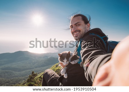 A man tourist with a cat makes a selfie in the mountains at dawn. Young handsome guy with his cat with camping equipment joyfully greet the morning sun at the top of the mountain. 