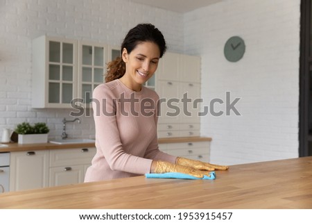 Millennial latin housewife enjoy easy putting modern designed kitchen in order wipe dust from wooden counter. Professional cleaning lady in rubber gloves remove dirt from work top using soft duster Royalty-Free Stock Photo #1953915457