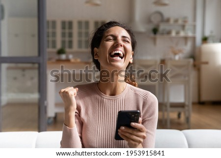 Winner time. Overjoyed young latina female sit on sofa alone hold phone laugh raise up fist proud with victory in game lottery online. Happy excited woman celebrate success scream yes full of emotions Royalty-Free Stock Photo #1953915841