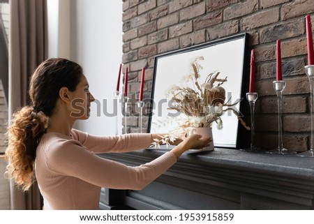 Family home. Happy hispanic female wife make rearrangement in living room place bunch of dry flowers on fireplace to get cozy apartment. Young female renter tenant decorating new house after moving in