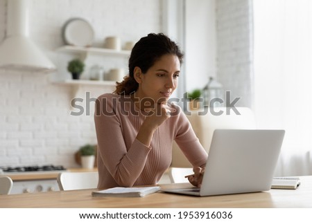 Web lecture. Focused young latina female sit at kitchen table look at pc screen listen to teacher think on problem to solve. Attentive lady remote student learn from home watch webinar training online Royalty-Free Stock Photo #1953916036