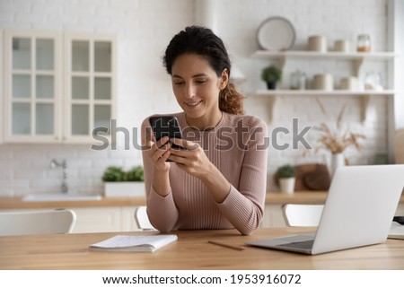 Confident latin female remote employee freelancer sit at kitchen table search files on phone to forward on laptop via home wireless network. Smart young lady user sync restore data using two gadgets