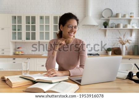 Smiling young latin lady learn remotely from home look on laptop screen distracted from reading paper literature. Positive woman student do academic research study printed books using online library