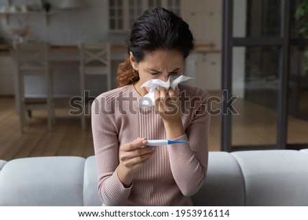 Having fever. Young latin female sit on couch at home feel sick bad sneeze blow nose to paper napkin look on thermometer display. Ill millennial woman measure temperature feel upset to see high degree Royalty-Free Stock Photo #1953916114
