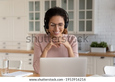 Young hispanic lady customer support manager on remote work wear headphone look on pc screen talk to client help offer advice consultation. Latin woman trainee listen to online webinar at home kitchen