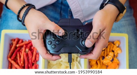 Young man holding a black game controller while eating snacks, video games and entertainment concept