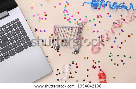 Laptop and Shopping trolley with Colored streamer and confetti on beige background. Holiday shopping, sale