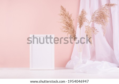 Soft pastel pink interior with blank photo frame, white silk curtain and beige fluffy reeds on wood shelf, copy space.