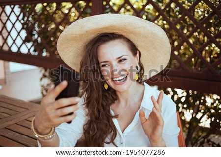 happy modern 40 years old woman in white shirt with hat having webcam meeting on a smartphone in the terrace of guest house hotel.