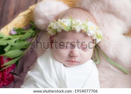 Baby girl in a wicker basket of vine decorated with burgundy peonies in a light winding and a flower wreath on her head. Spring photo. Flowers and children. Happy motherhood 