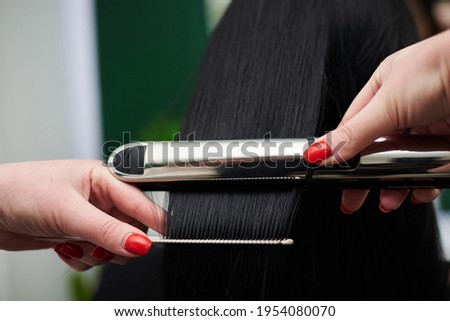 Young brunette woman getting ready for party. Professional hairdresser making hairstyling for female client with hair straightener in front of mirror. Close-up picture of work process in barber shop.
