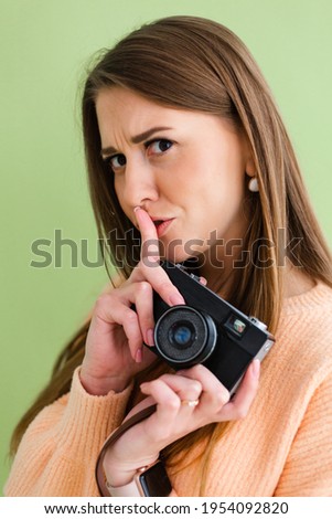 Pretty european woman in casual peach sweater isolated on green  background with camera in hands positive shows shh sign with finger, silence gesture 