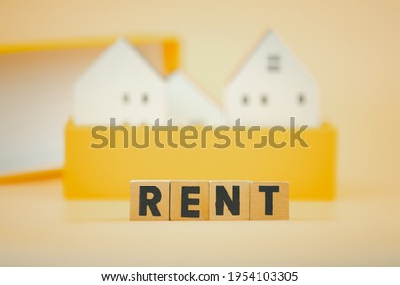 Wooden cubes with the text RENT with against background of small white home in yellow box. Renting property concept.