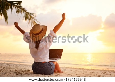 Work from anywhere. Rear view of young woman, female freelancer in straw hat working on laptop, keeping arms raised and cellebrating success while sitting on the tropical sandy beach at sunset Royalty-Free Stock Photo #1954135648