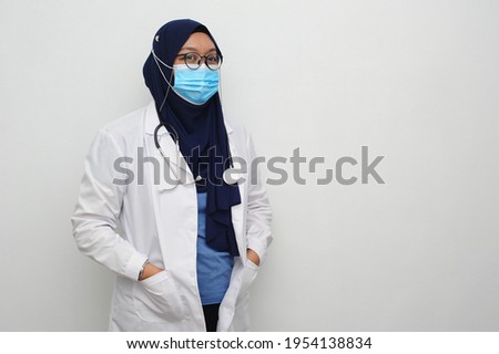 Asian female doctor wearing hijab standing with hand in pocket white coat with hanging stethoscope in white background with copy-space.

