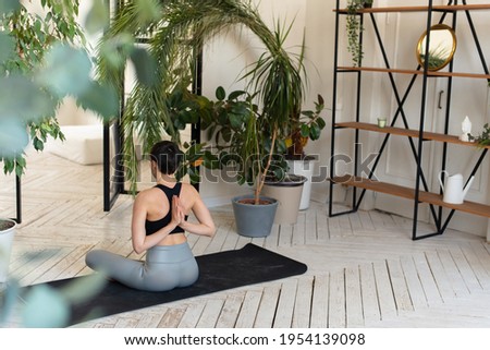 Young darkhair woman practicing yoga in the morning at her home near plants. The woman is engaged in self-determination, doing yoga exercises Royalty-Free Stock Photo #1954139098