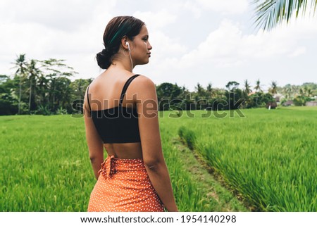 Back view of fit young ethnic brunette in crop top enjoying music in earphones and admiring nature in tropical garden during summer holidays