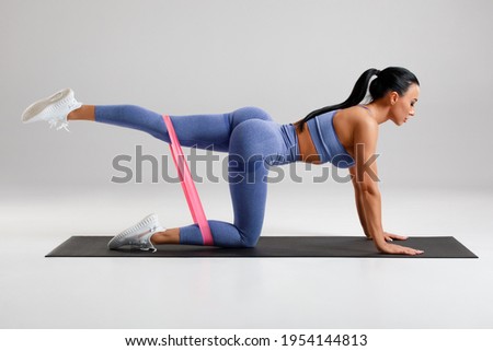 Fitness woman doing kickback exercise for glutes with resistance band on gray background. Athletic girl working out donkey kicks Royalty-Free Stock Photo #1954144813