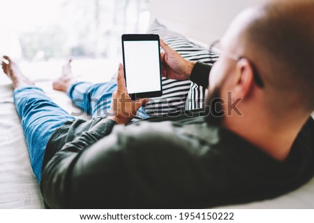 Back view of crop unshaven male with eyeglasses spending time on sofa browsing tablet and checking social network during break