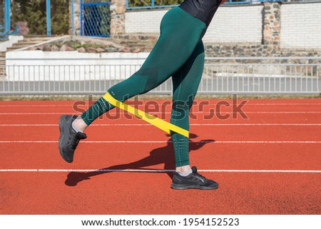 Fitness and sport concept, outdoor workout. Woman's legs work out with the help of a fitness elastic band on the red track.