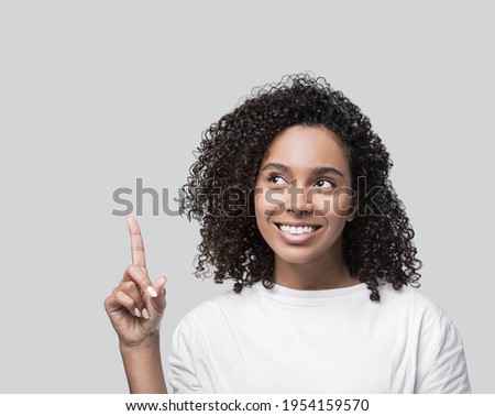Studio portrait of a beautiful young woman pointing up Mixed race girl wearing white t-shirt showing product. Isolated on grey background. People, lifestyle, beauty, advertisement concept