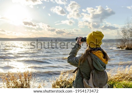 Young woman looking through binoculars at birds on the lake. Birdwatching, zoology, ecology. Research in nature, observation of animals. Ornithology Royalty-Free Stock Photo #1954173472