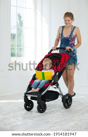 Caucasian young woman and toddler son in stroller indoors. Boy holding book and looking at pictures.